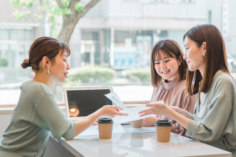 カフェで3人の女性が楽しそうに会話をしながら書類を見ている。